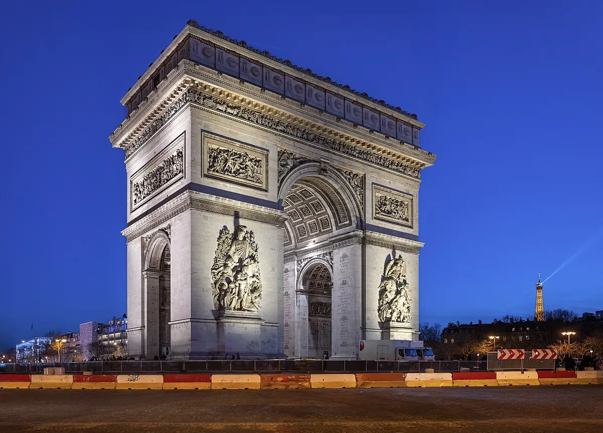 The Arc de Triomphe lit against a deep blue evening sky, the Eiffel Tower small in the distance