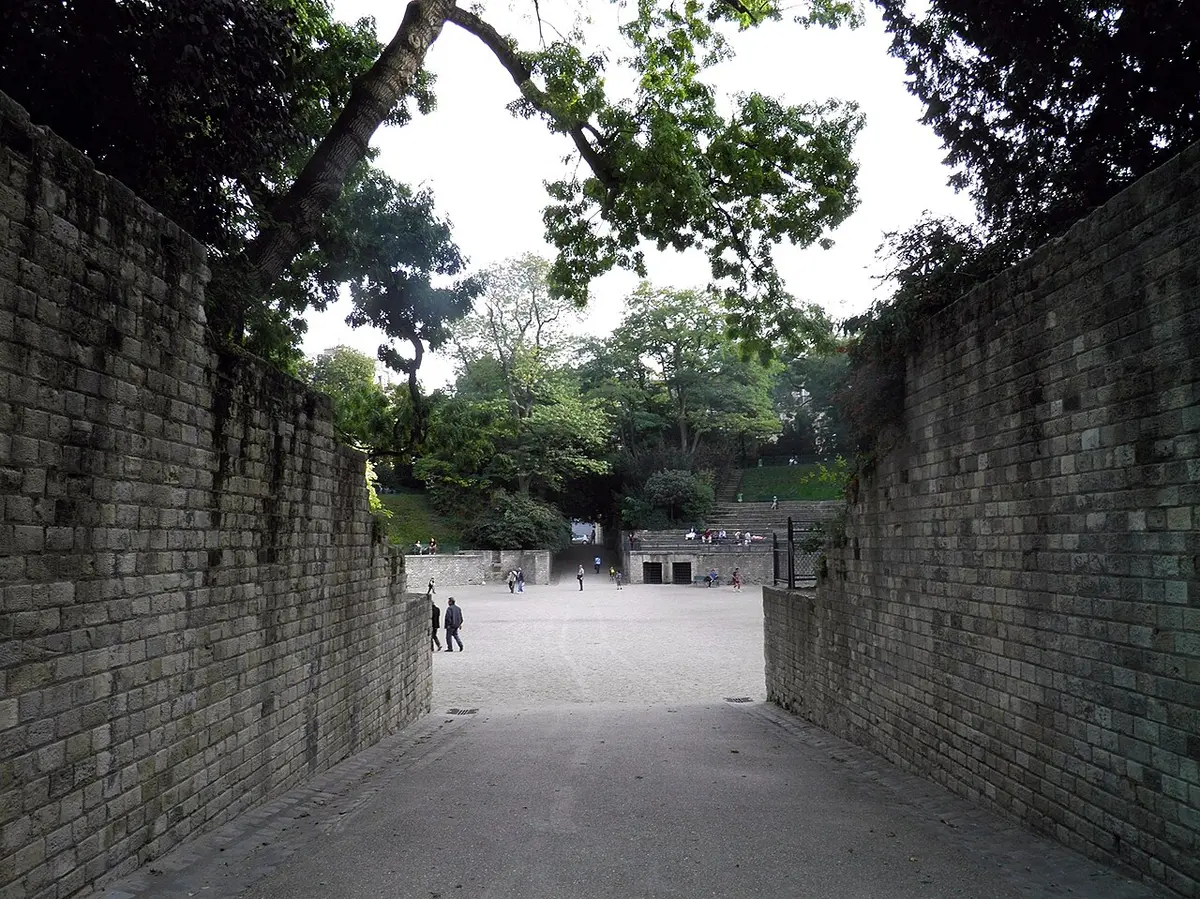 A passage between old stone walls leading into the sandy floor and stepped seats of the Arènes de Lutèce