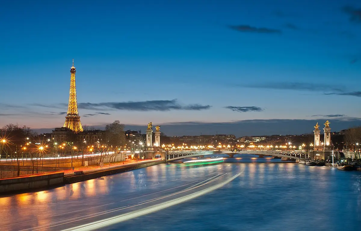 The Eiffel Tower lit at dusk beyond the Pont Alexandre III, with light trails on the Seine