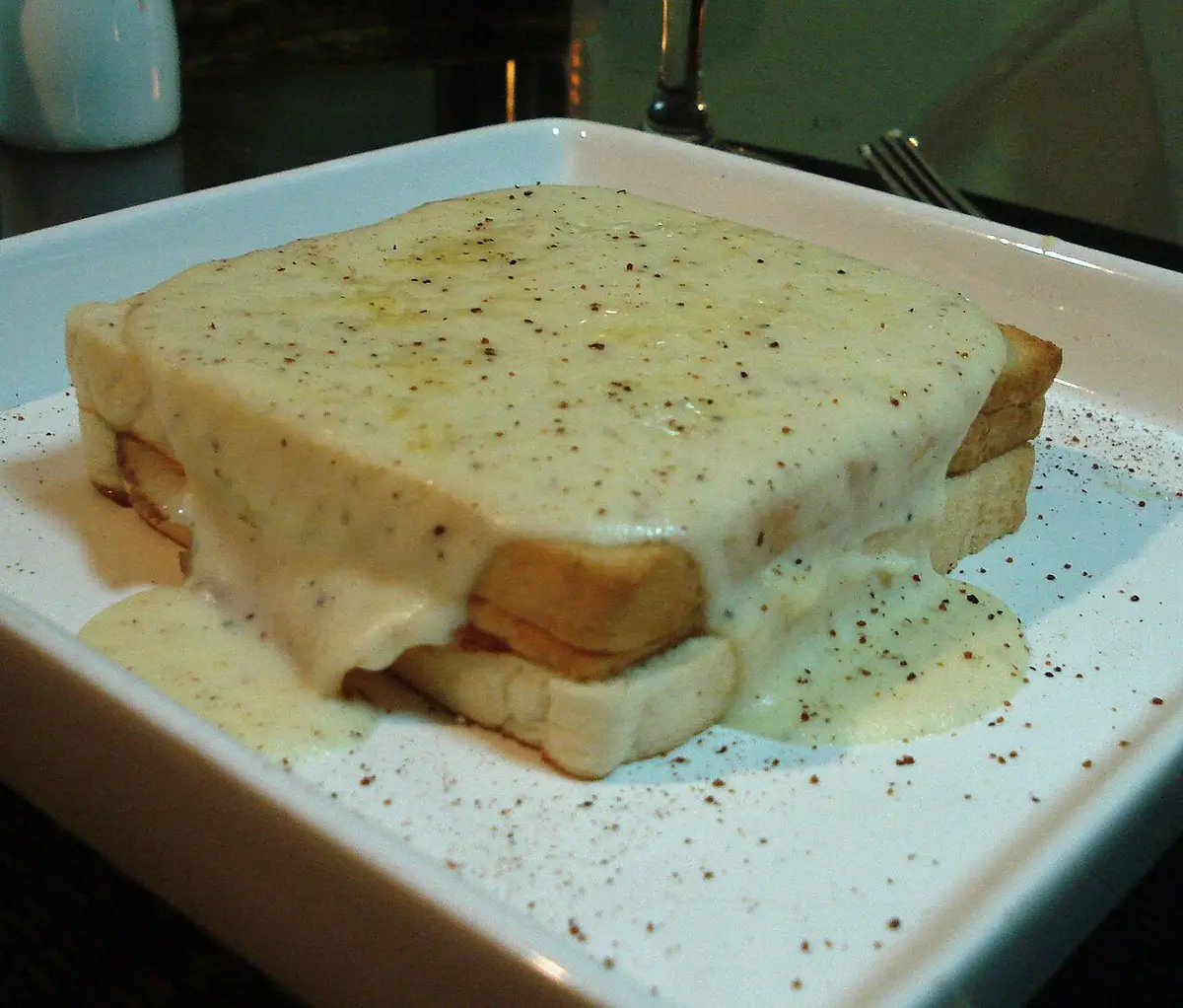 A croque-monsieur covered in melted béchamel and cheese on a square plate