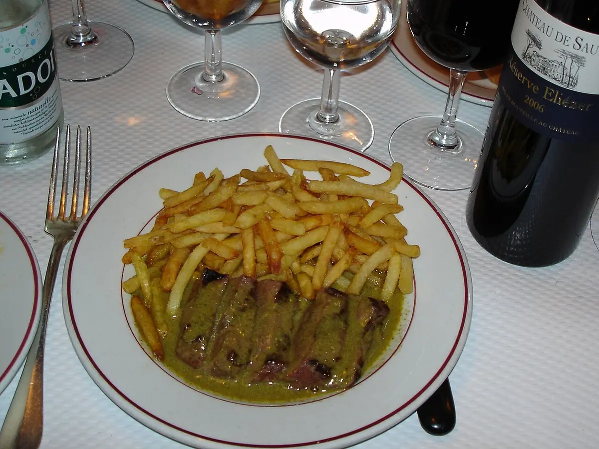 Sliced steak in a green butter sauce under a pile of thin fries on a bistro plate