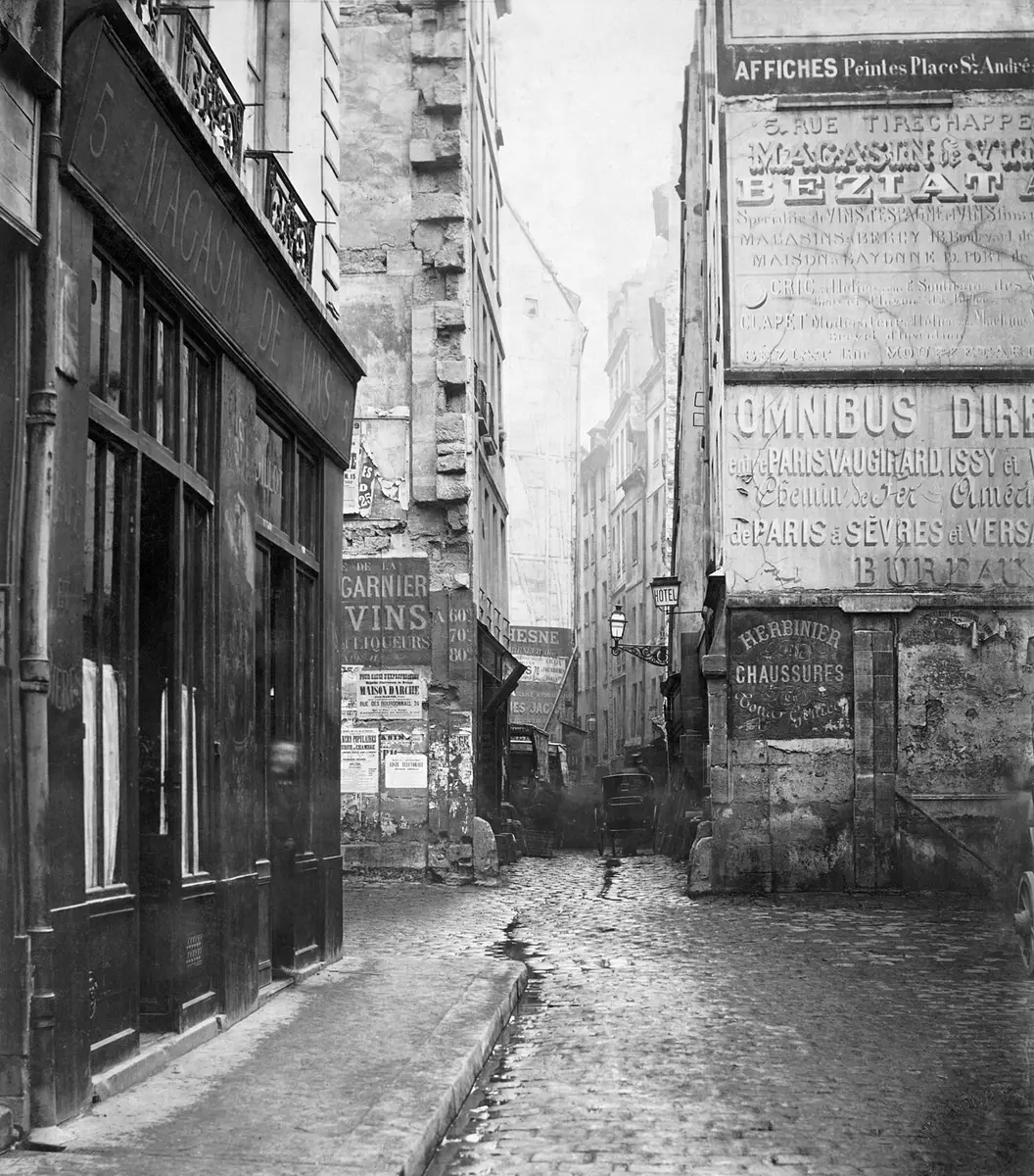 Black and white photograph of a narrow cobbled street between tall old houses, with painted signs on the walls