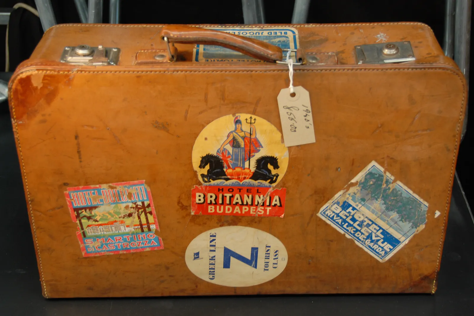 An old leather suitcase covered in vintage hotel and shipping-line luggage labels, a paper tag hanging from the handle