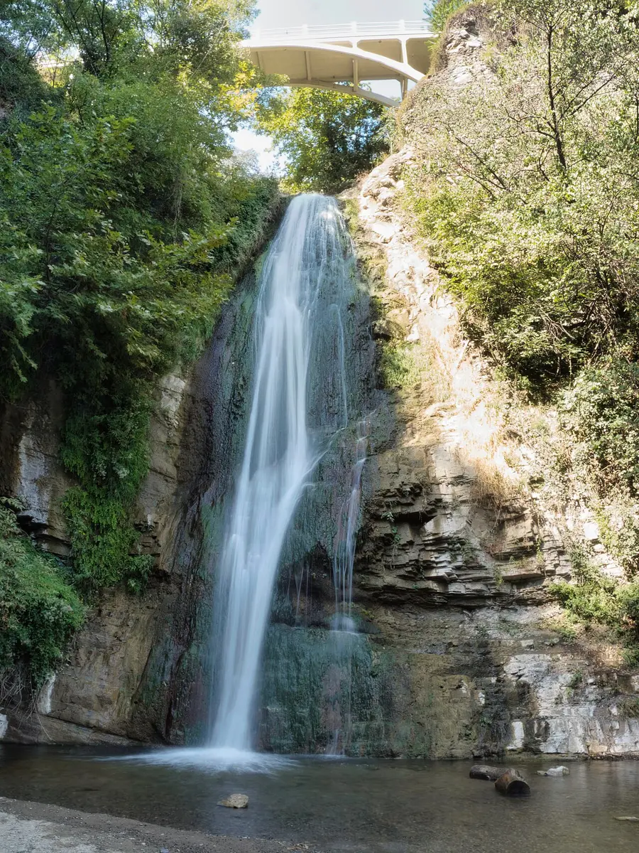 A waterfall dropping into a pool in a rocky gorge, with a white arched bridge high above