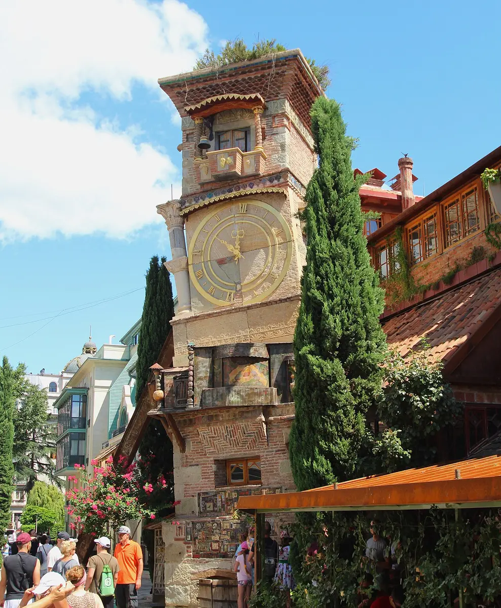 The leaning clock tower of the Gabriadze puppet theater, faced with tiles, beside a cypress