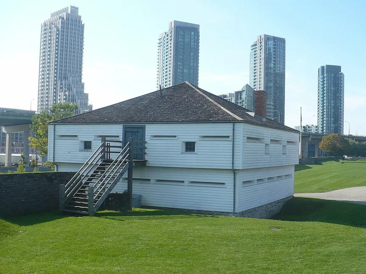 A white wooden blockhouse on a grassy earthwork at Fort York, condo towers rising behind
