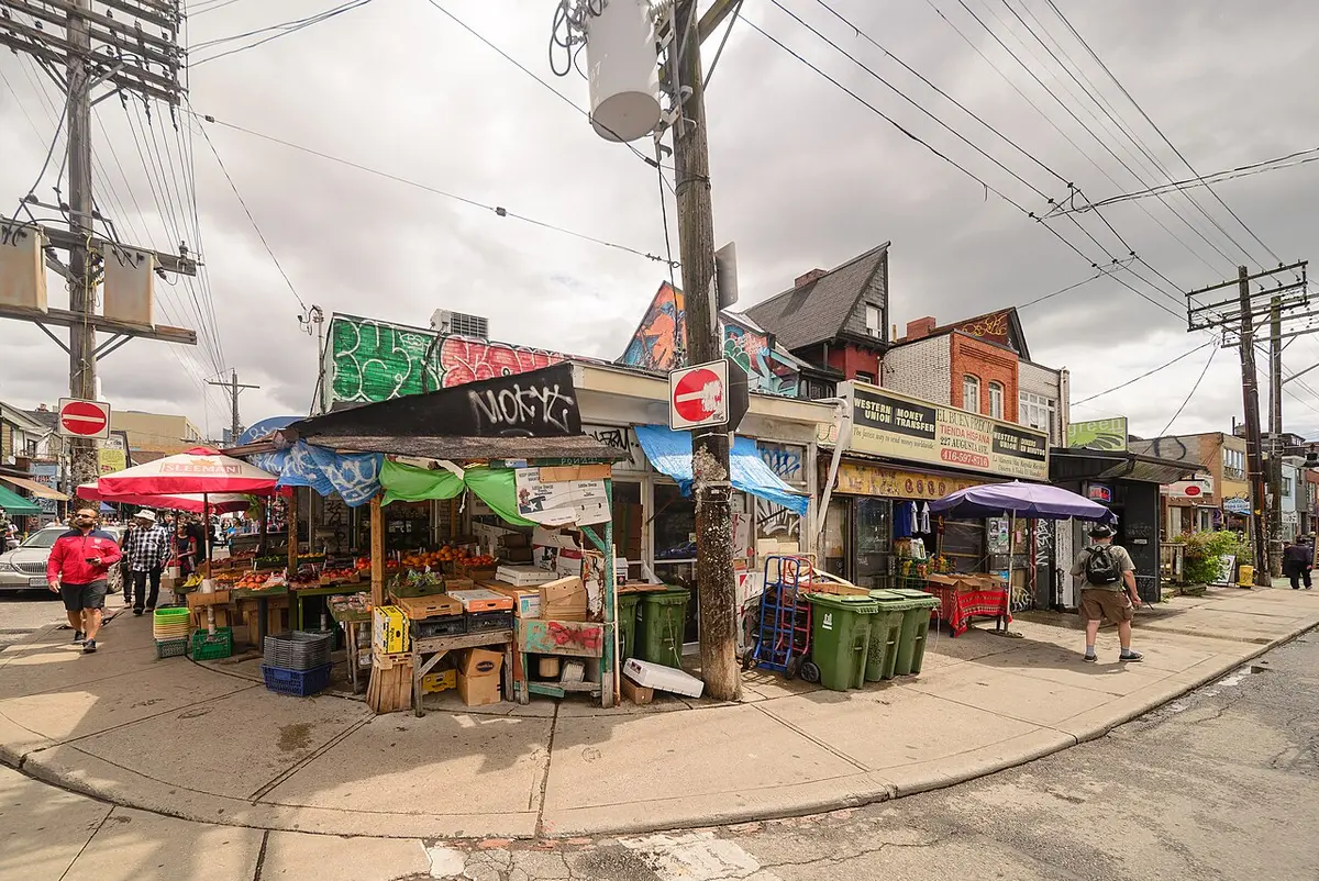 Fruit and vegetable stalls under awnings at a street corner in Kensington Market, colorful houses behind