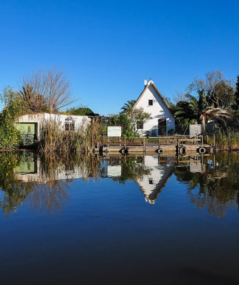 A whitewashed barraca with a steep roof and a cross on top, beside a jetty on a canal, reflected in the water