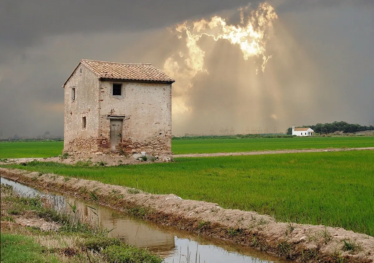 An old stone farm hut beside a canal among green rice fields under a stormy sky