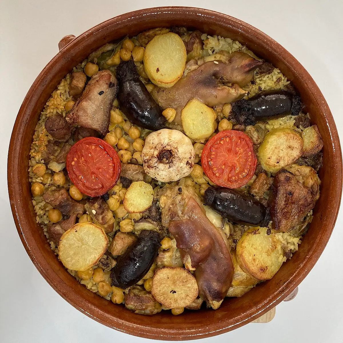 Arròs al forn in a clay dish, with chickpeas, black pudding, pork, potato, tomato and a head of garlic
