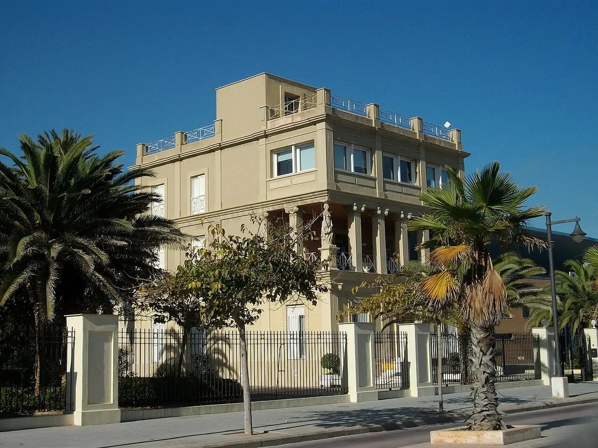 A cream-colored villa with columns and a roof terrace behind palm trees, on the seafront